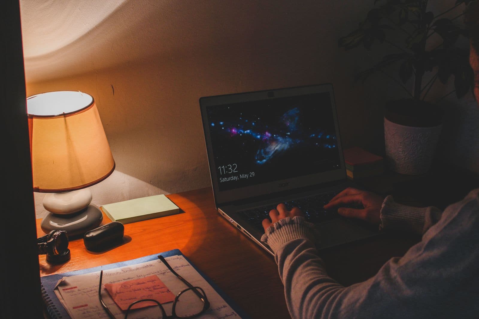 A desk at night with a warm lamp, laptop and notes
